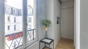 Un couloir étroit avec une grande fenêtre, une petite table sur laquelle est posée une plante, et une porte qui donne sur une autre pièce.