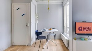 Une pièce avec une porte blanche, une table ronde avec deux chaises, un téléviseur affichant le message « CHAMBRE PRÊTE POUR L’ARRIVÉE DES CLIENTS » et une fenêtre avec des stores.