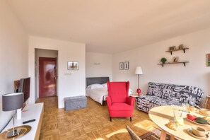 Ett modernt hotellrum med en säng, en röd fåtölj, en mönstrad soffa, ett matbord med frukosttillbehör och en tv.