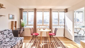 A modern living room with a sofa, a dining table with chairs, large windows with curtains, and a view of the cityscape. - GuestReady - Magnificent Parisian Apartment (Paris)