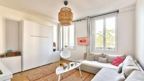 A modern, well-lit living room with a sofa, a coffee table, a hanging light fixture, and a window with a view. - GuestReady - lovely stay near Moulin Rouge (Paris)