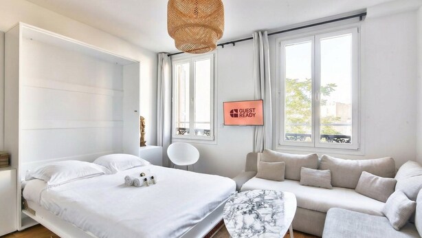 A modern hotel room with a bed, a sofa, a TV displaying "GUEST READY", and large windows. - GuestReady - lovely stay near Moulin Rouge (Paris)