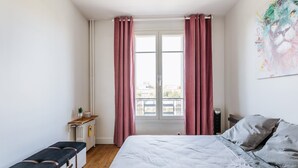 A bedroom with a bed, a bench, a radiator, a window with curtains, and a painting on the wall.