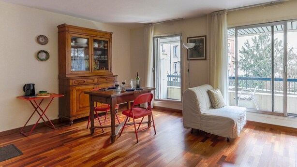 Een kamer met een houten eettafel, rode stoelen, een witte bank, een houten kast en een groot raam met uitzicht op gebouwen en bomen.