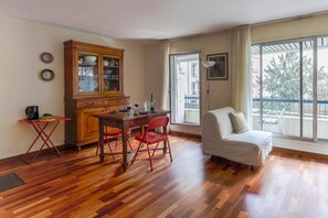 Une pièce comprenant une table à manger en bois, des chaises rouges, un canapé blanc, un meuble en bois et une grande fenêtre donnant sur des immeubles et des arbres.