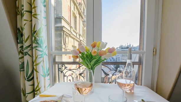 Un tavolo da pranzo con piatti di cibo, bicchieri di vino e un vaso di tulipani su un balcone con vista sulla città.