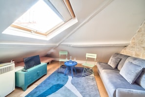 Una habitación compacta en el ático, con techo inclinado, televisor sobre un mueble verde azulado, sofá gris, mesita con silla y tragaluz.
