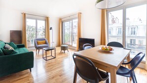 A modern living room with a sofa, a dining table with chairs, a floor lamp, and large windows offering a view of the city. - GuestReady - Superb Stay in the Heart of Paris (Paris)