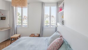 Ein Schlafzimmer mit Bett, kleinem Tisch, einer Bücherablage und einem Fenster mit Blick auf Gebäude.