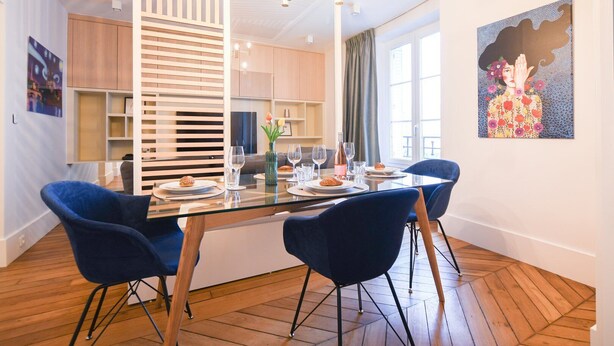 A modern dining area with a glass table, blue chairs, and a decorative painting on the wall.