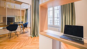 A modern room with a wooden herringbone floor, a desk with a laptop, a dining area with chairs, and a large window with curtains.
