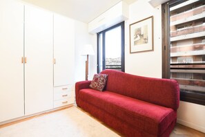 Um sofá vermelho com uma almofada estampada, um guarda-roupa branco, um abajur e uma janela com vista para a cidade.