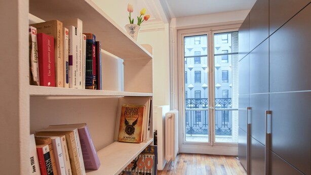 A narrow hallway with a bookshelf filled with books, a window with a view of buildings, and a modern kitchen area with cabinets. - GuestReady - Bright Apt in the new Quartier Latin (Paris)