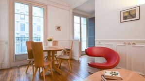 A dining area with a wooden table and chairs, a red chair, and a window with a view of buildings. - GuestReady - Bright Apt in the new Quartier Latin (Paris)