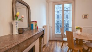 A room with a marble fireplace, a dining table with chairs, a vase with flowers, a mirror, and a window with a view of buildings. - GuestReady - Bright Apt in the new Quartier Latin (Paris)