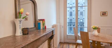 A room with a marble fireplace, a dining table with chairs, a vase with flowers, a mirror, and a window with a view of buildings.