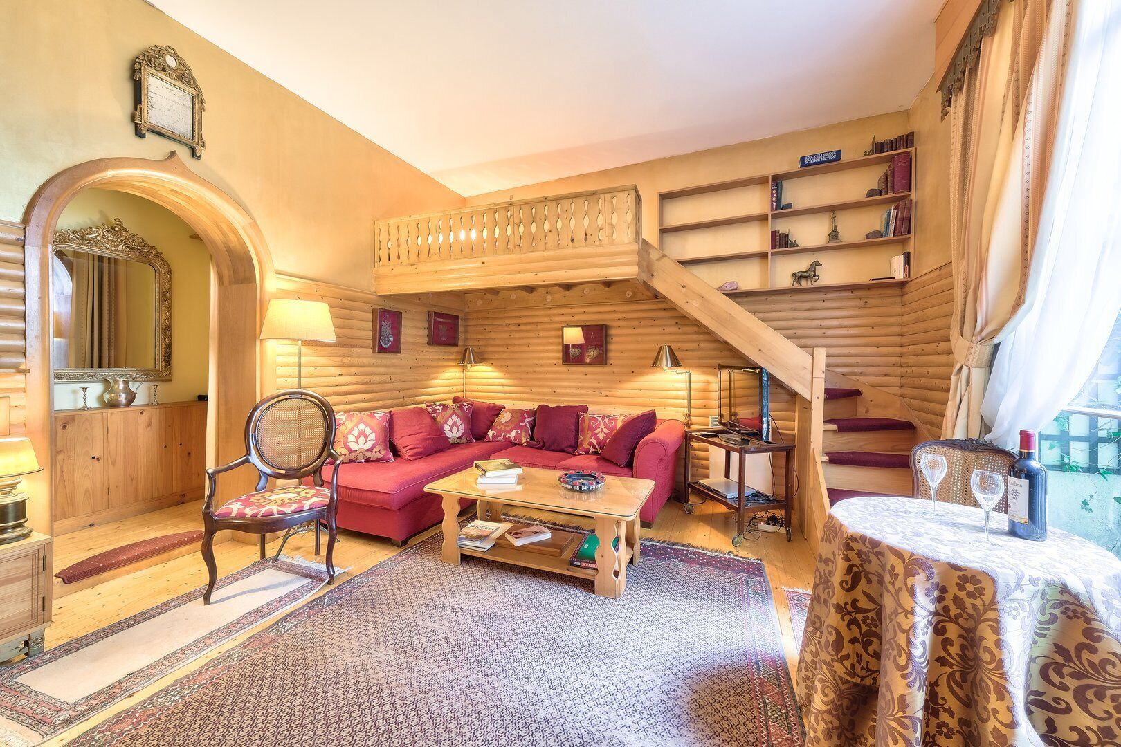 A cozy log cabin interior with a red sofa, wooden furniture, a staircase, and a bookshelf.