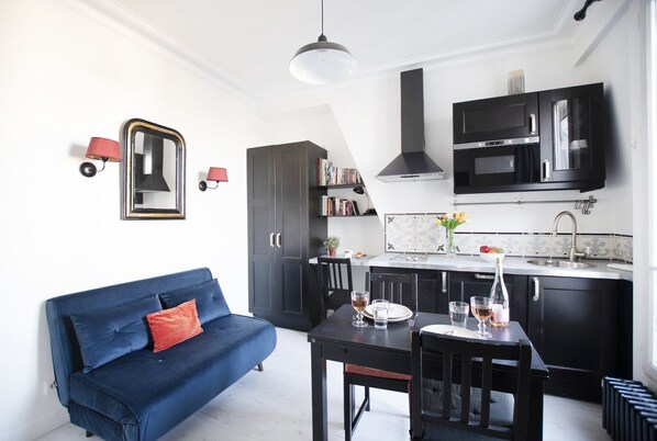 A compact kitchen with black cabinets, a dining table, and a blue sofa. - GuestReady - Cozy and Bright with Notre Dame View (Paris)
