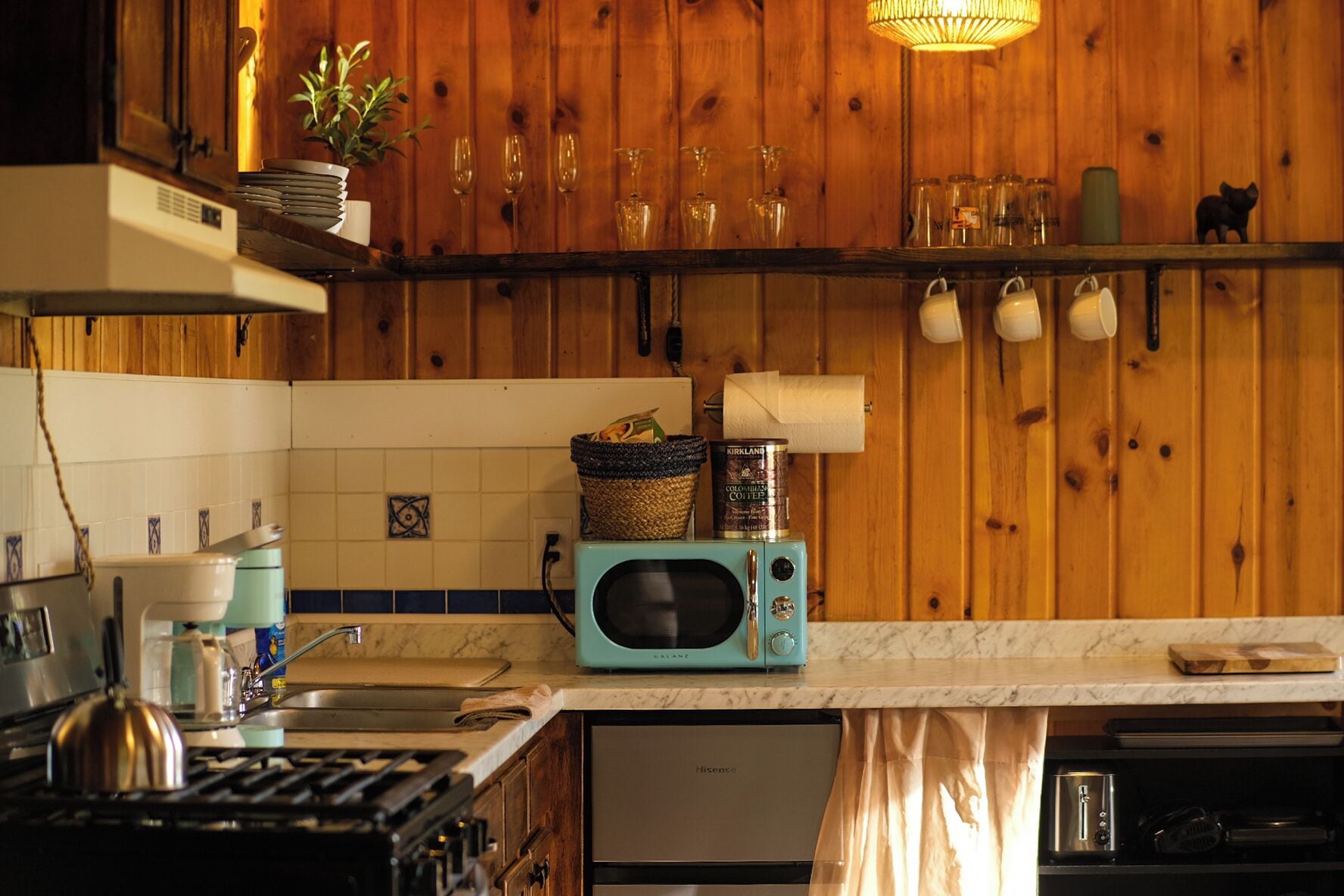 Een keuken met houten lambrisering, een blauwgroene magnetron, een gasfornuis en een gootsteen.