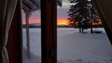 A view through a window with curtains, featuring a snowy landscape, a tree, and a sunset.