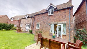 A brick house with a patio, a wooden table, and chairs. - Stunning Coastal Cottage - Sleeping 5 in 2 Bedrooms - Pool and Beach Access Included. (Filey)