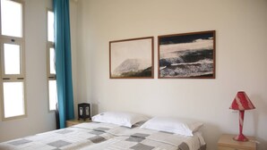 Ein Schlafzimmer mit einem Bett, zwei gerahmten Bildern, einem Nachttisch, einer Lampe und einem Fenster mit Vorhängen.