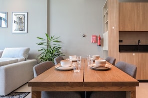 A modern dining area with a wooden table set for four, surrounded by grey upholstered chairs. A potted plant is on the left, and a kitchen area is visible to the right. - Luxurious 1BR Suite Signature Livings in Dubai (Dubai)