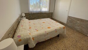 A bedroom with a bed, a nightstand, a window with curtains, a door, and a stone-patterned wall. - SAONA VILLAS (VN CARL) : Villa, private swiming pool, wifi, 4 people (Tres Calas)