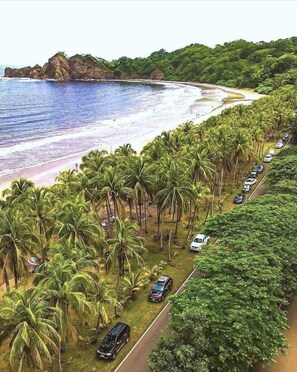 Uma estrada costeira bordeada por palmeiras e carros estacionados, que leva a uma praia com águas azul-claras e afloramentos rochosos.