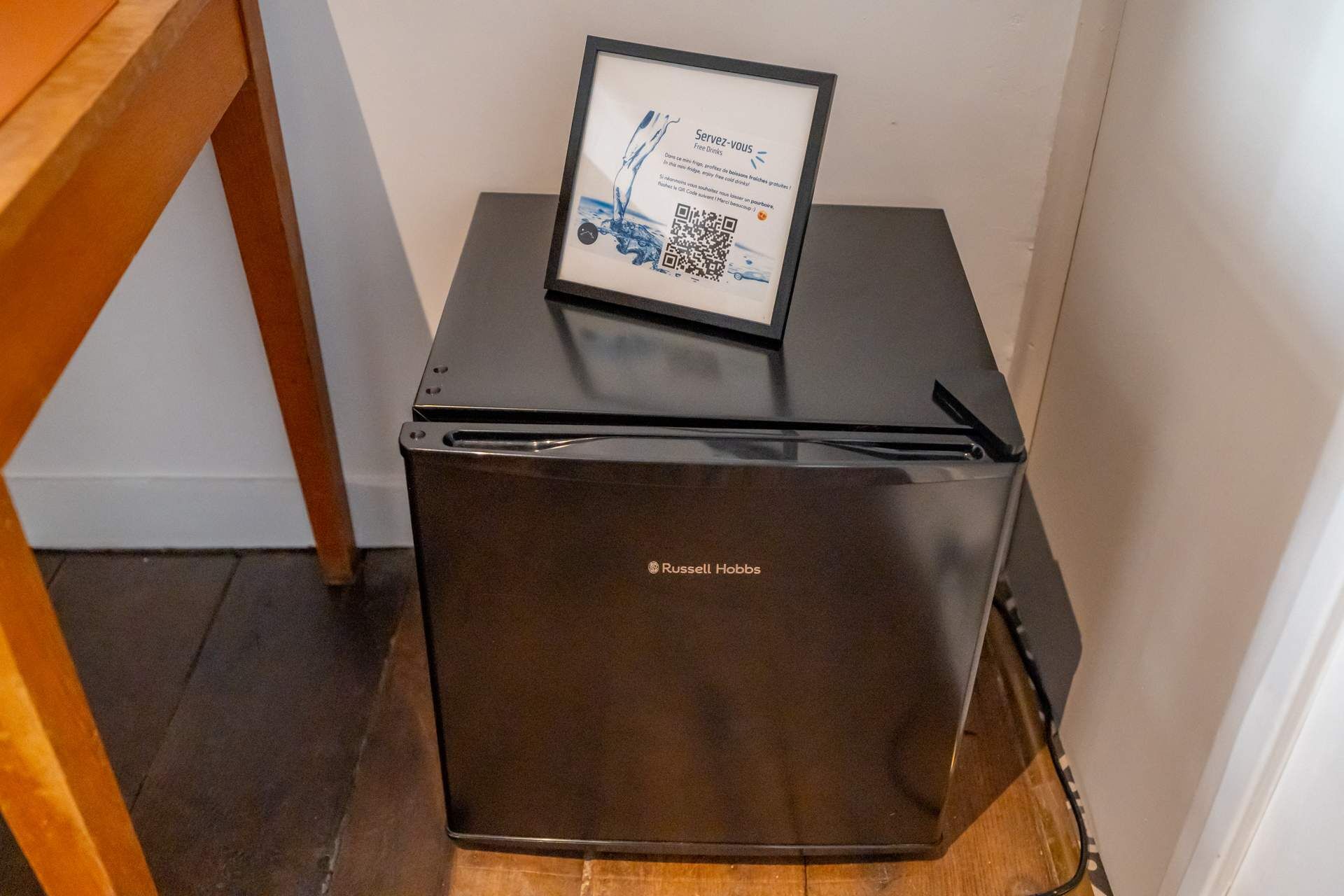 Un refrigerador compacto con un código QR y un letrero en la parte superior, ubicado en un rincón de una habitación con una silla de madera y una pared blanca.