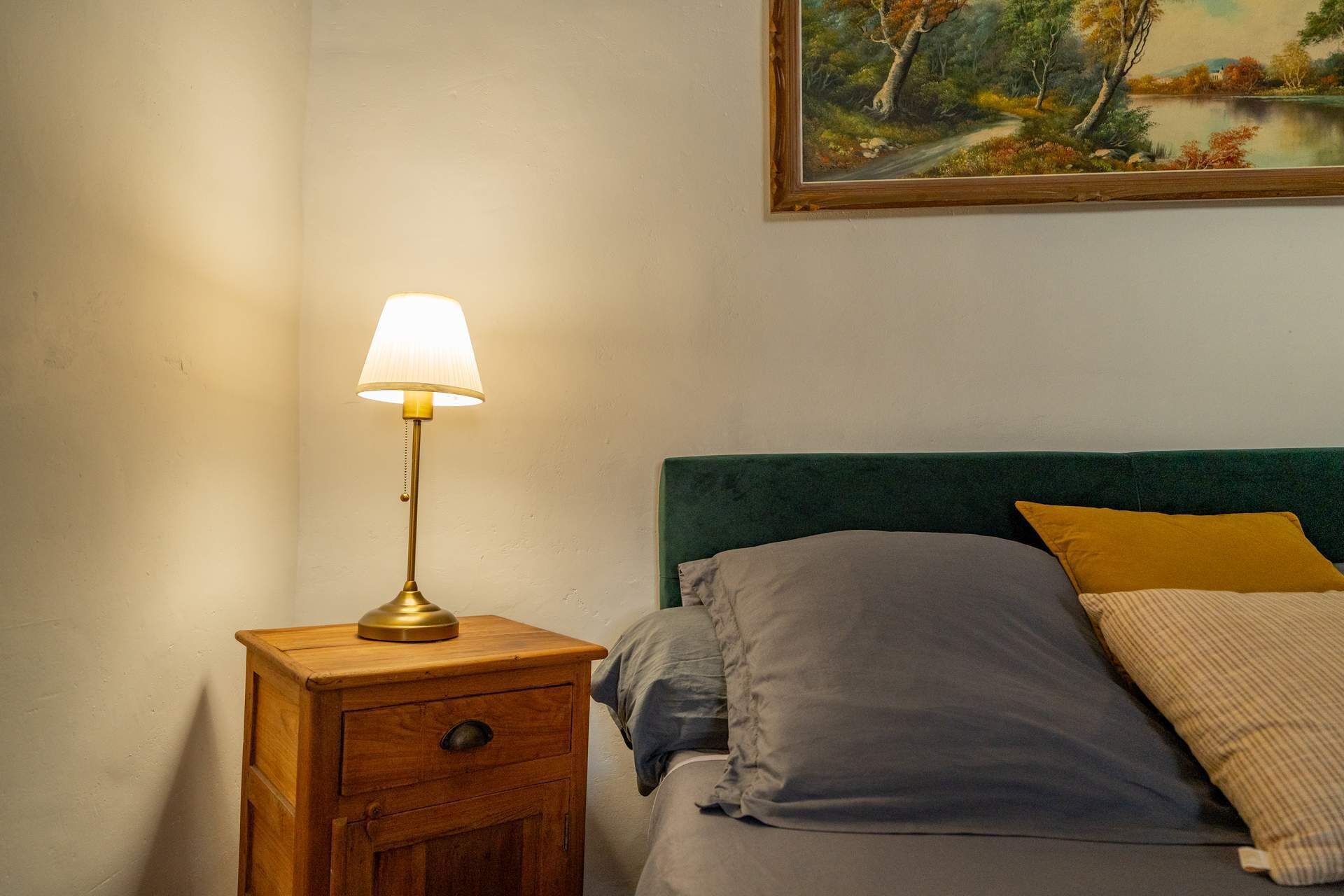 Et soveværelse med et trænatbord, en lampe, en seng med grøn gavl og et landskabsmaleri på væggen.