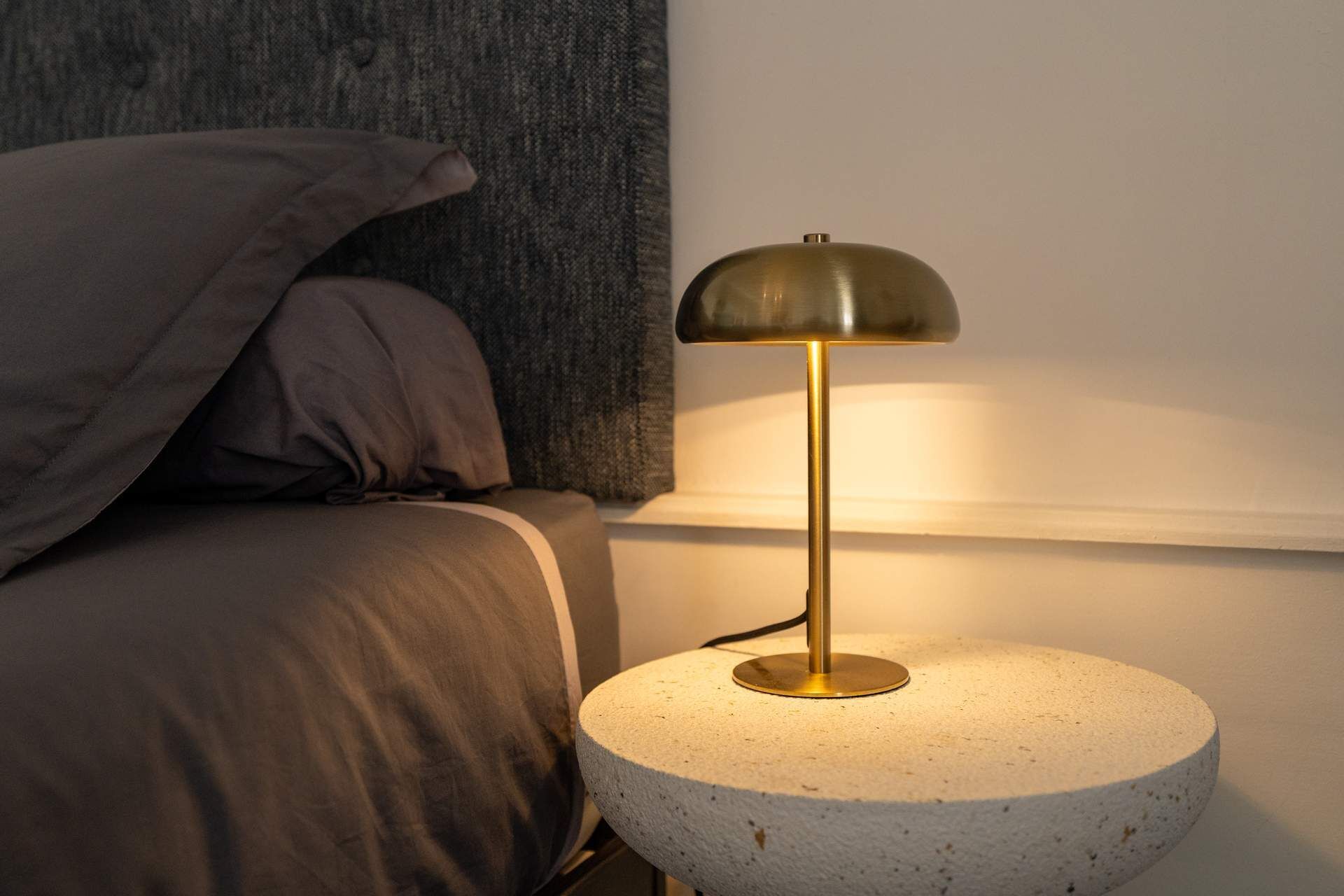 Een ronde, gestructureerde tafellamp met metalen voet en een koepelvormige kap, geplaatst op een witte ronde tafel naast een bed met een grijze hoofdbord en kussens.