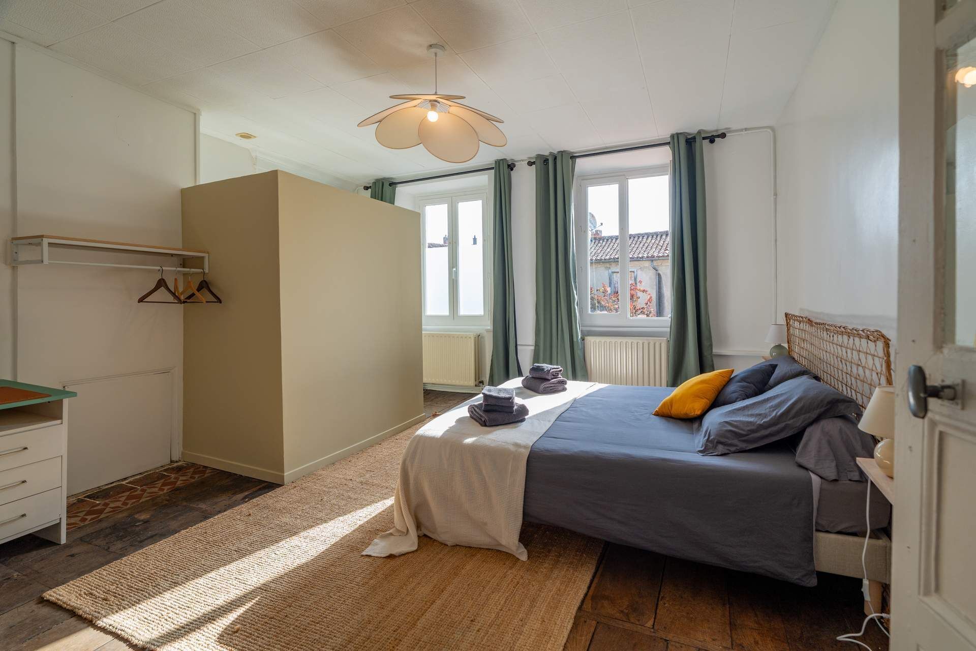 Un dormitorio con cama, una mesita de noche, una lámpara colgante, una ventana con cortinas y vistas a edificios.