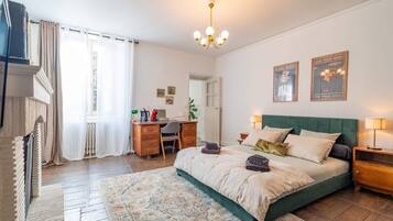 Un dormitorio con cama, escritorio, silla, radiador, ventana con cortinas y una lámpara de araña.