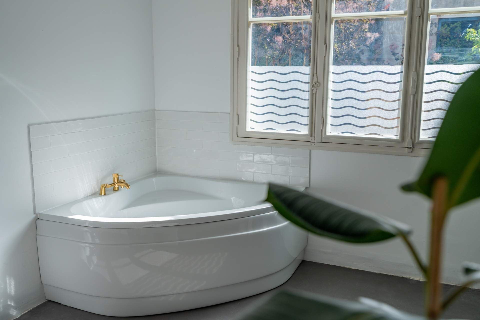 Eine Eckbadewanne mit goldfarbenem Wasserhahn, weißen Fliesenwänden und einem Fenster mit wellenmuster Blinds.