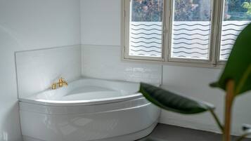 Eine Eckbadewanne mit goldfarbenem Wasserhahn, weißen Fliesenwänden und einem Fenster mit wellenmuster Blinds.