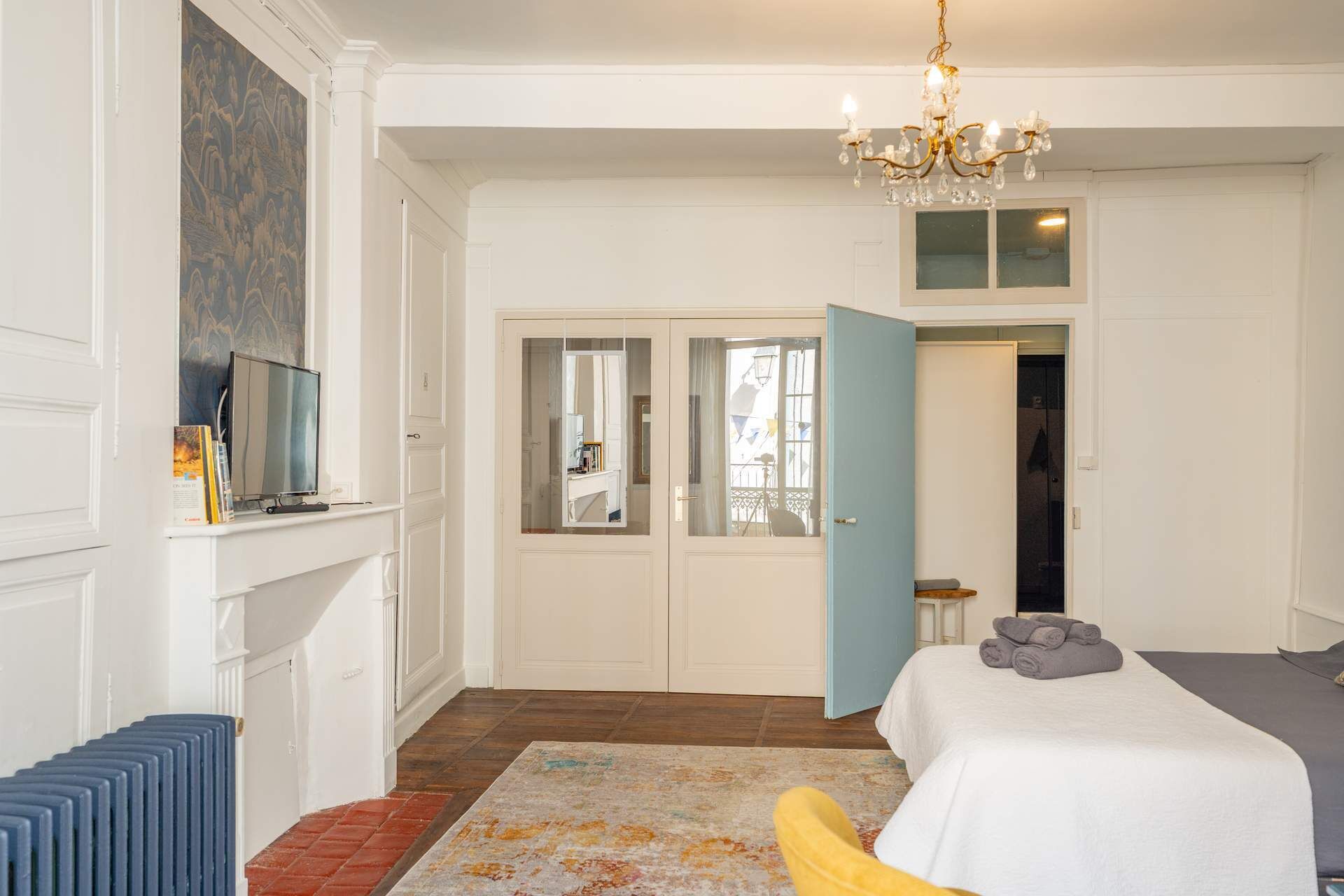 A bedroom with a bed, a chandelier, a mirror, a radiator, and a door leading to another room.