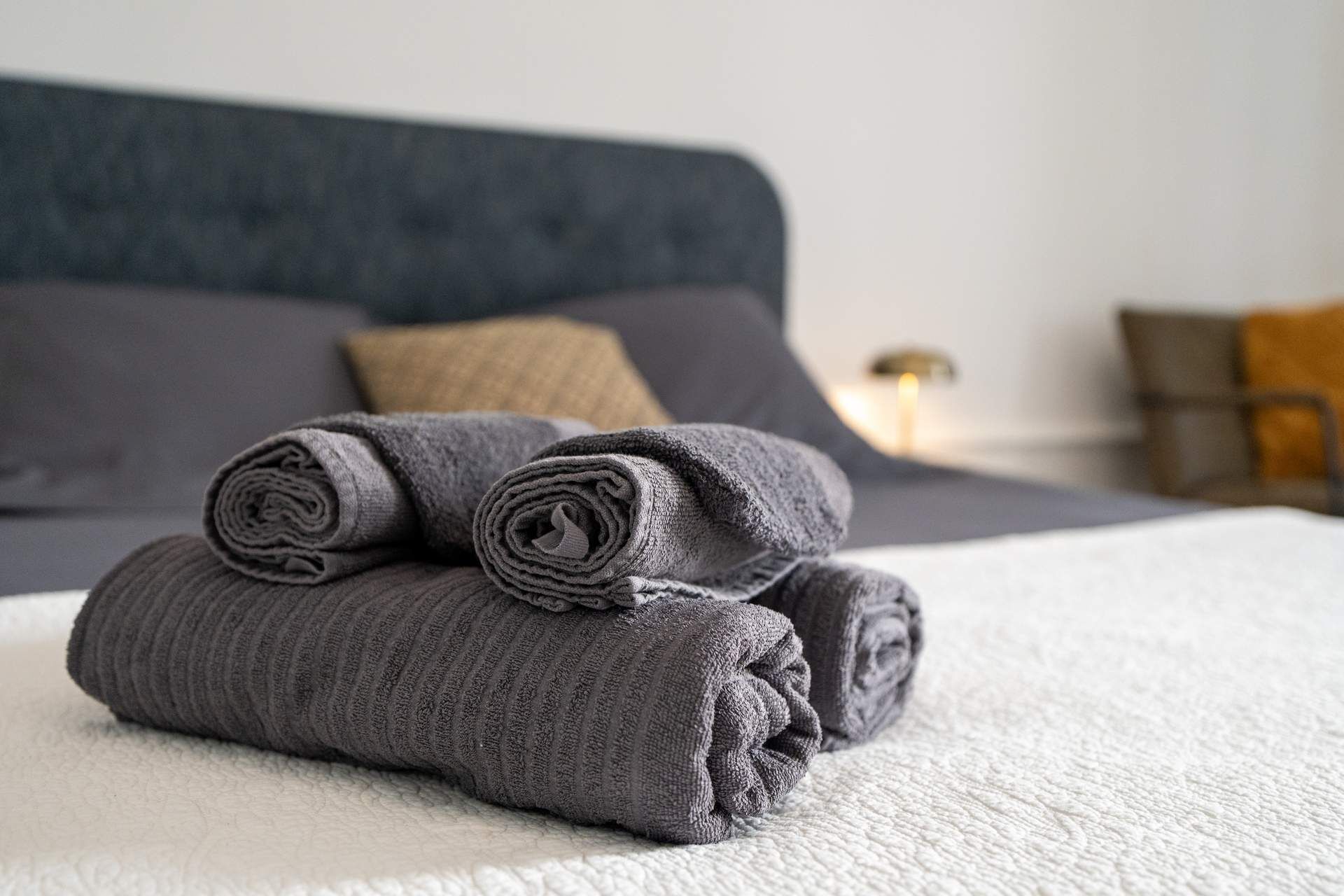 A neatly made bed with a grey blanket and rolled towels.