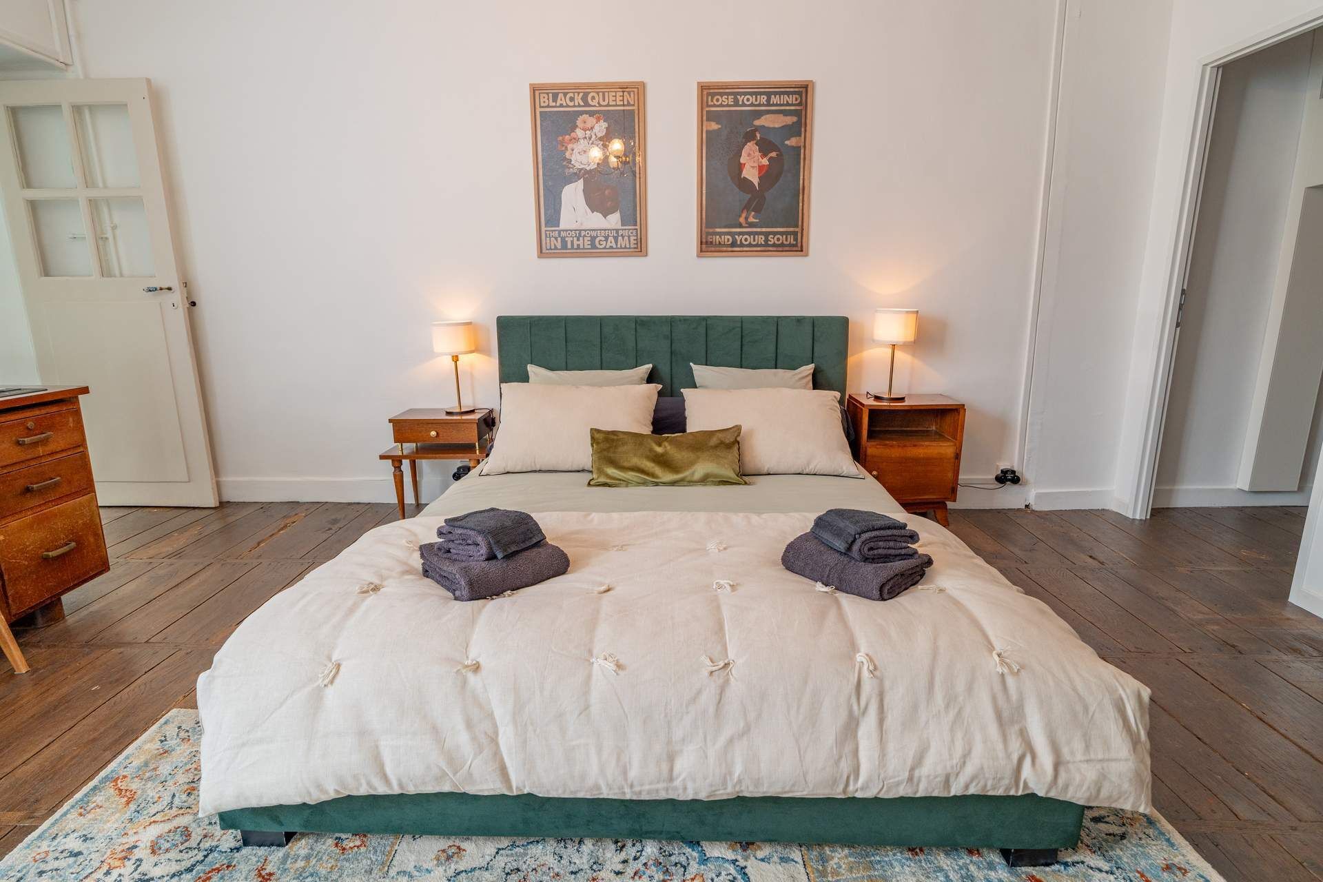 Een net opgemaakt bed met een groen hoofdeinde, twee nachtkastjes met lampen, en twee posters aan de muur.