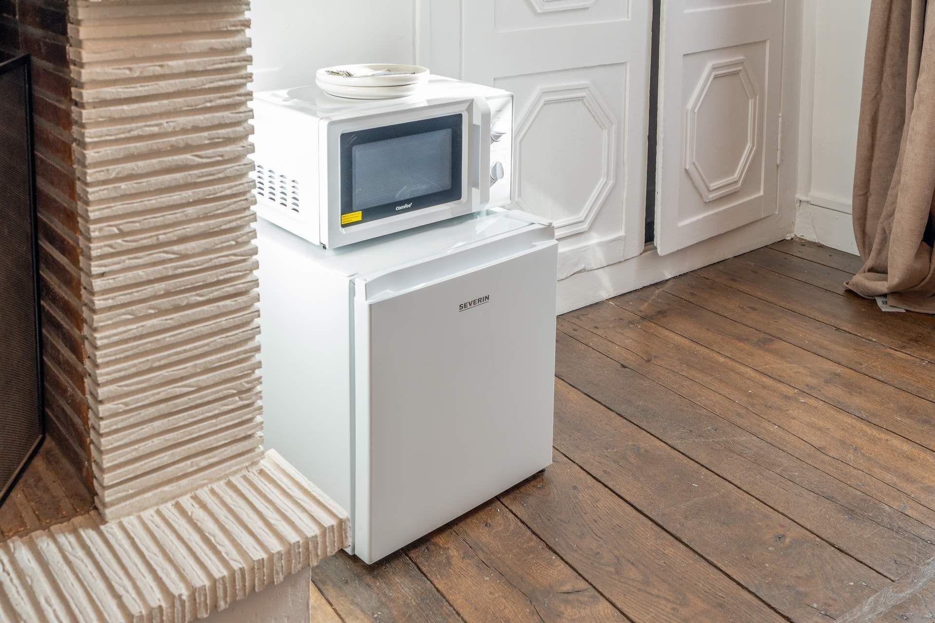 A compact white refrigerator with a built-in microwave on top, placed in a corner against a brick wall.