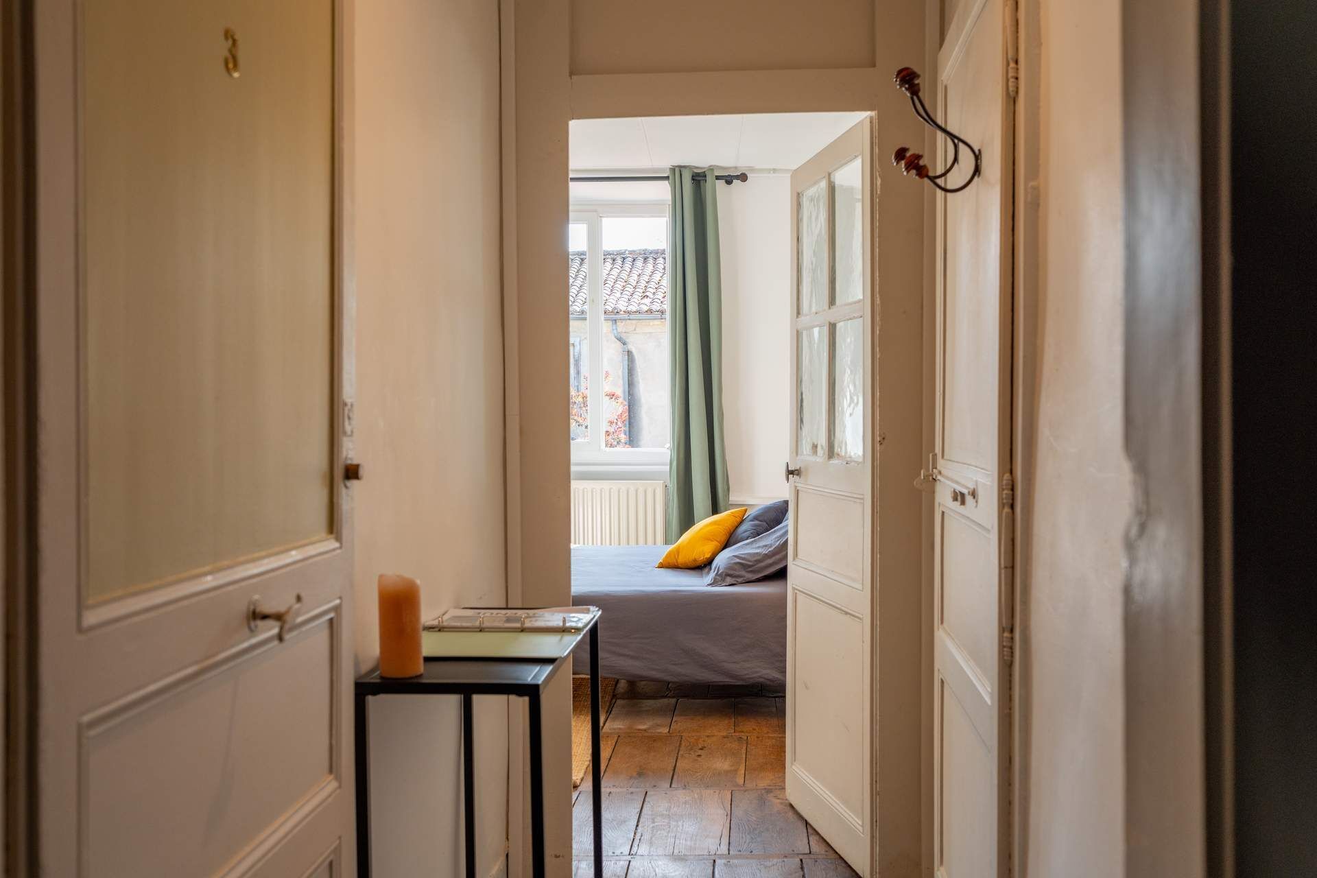 A narrow hallway leading to a bedroom with a bed, a bedside table, and a window with curtains.