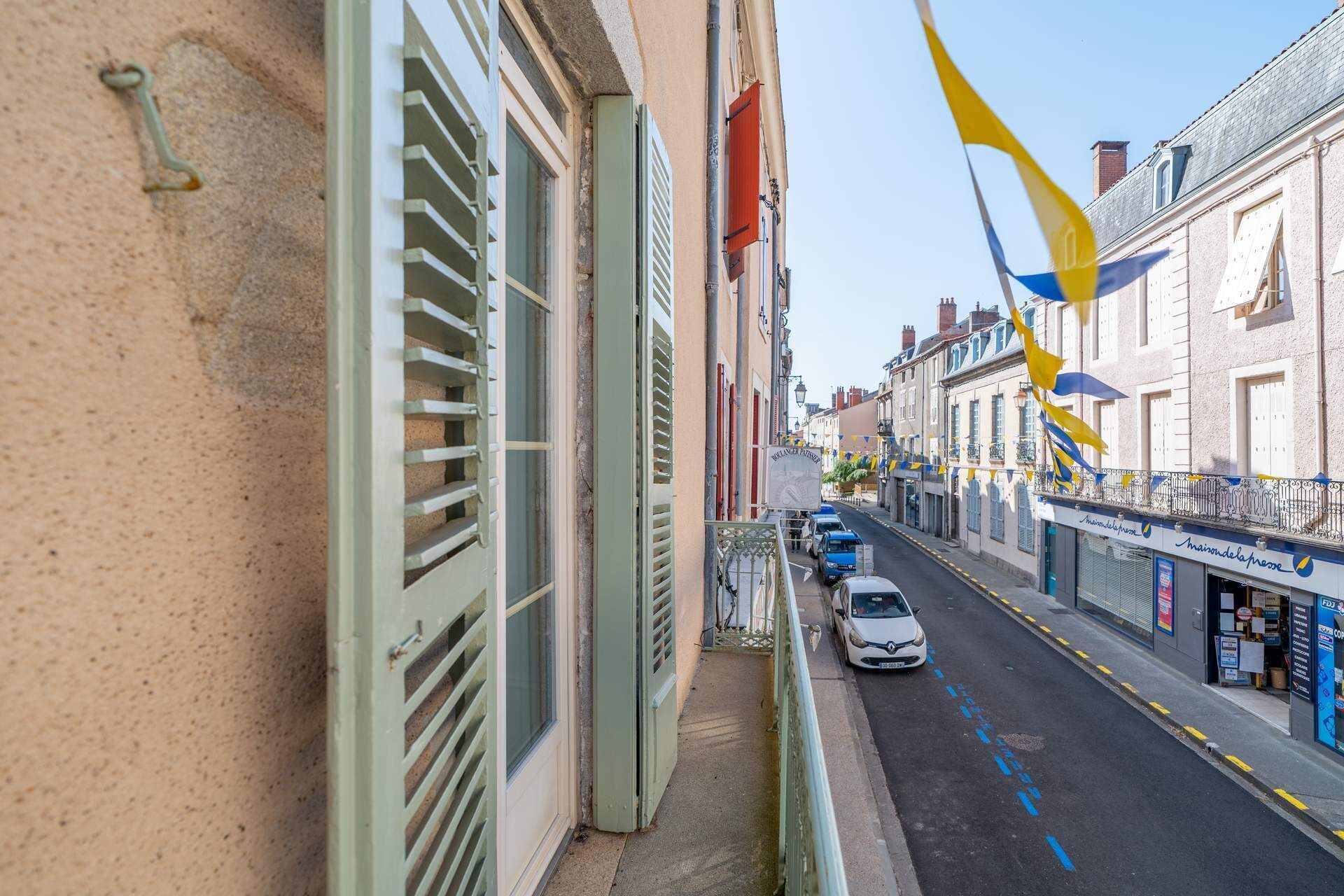 Une rue étroite, bordée de bâtiments des deux côtés, avec une voiture blanche garée sur le bord et des drapeaux jaunes et bleus flottant au-dessus des bâtiments.
