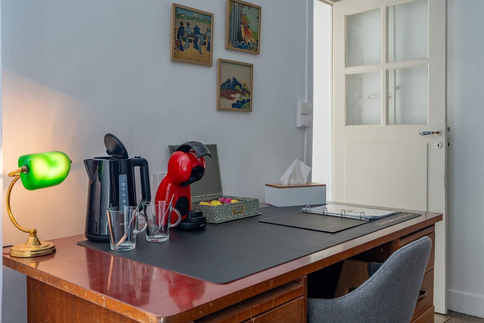 A wooden desk with a black coffee maker, a red electric kettle, a green desk lamp, and a tissue box. There are three framed artworks on the wall.