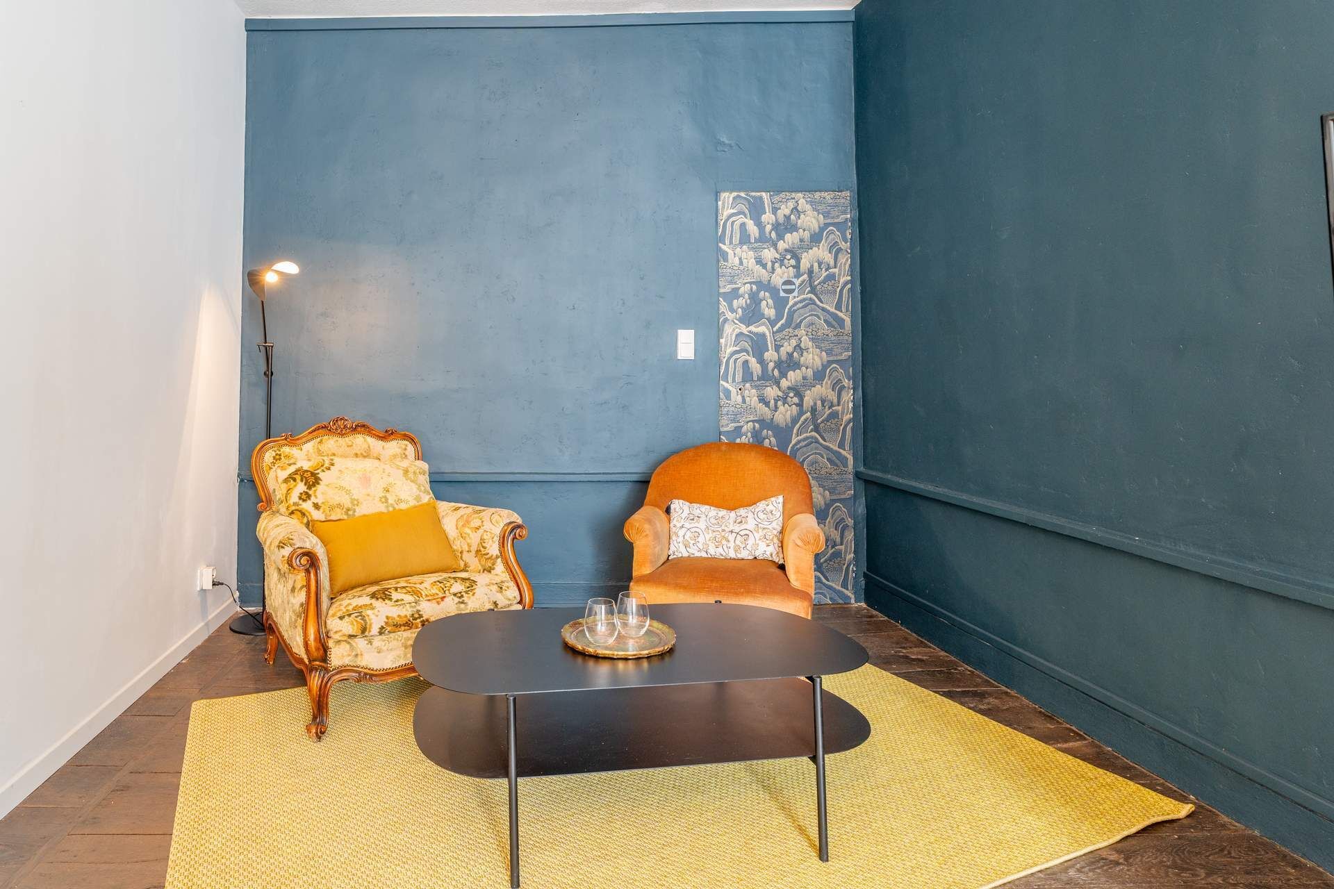 A room with a blue accent wall, a patterned tapestry, a vintage armchair with a yellow cushion, a modern armchair with a patterned cushion, and a small round table with a coaster and glasses.