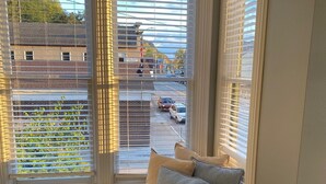 Una habitación con un banco empotrado debajo de una ventana con persianas blancas, vista a la calle y suelo de madera.