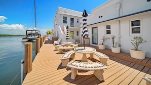 A wooden deck with outdoor seating and potted plants, a white building with a door and windows, and a sailboat docked nearby.