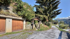 En smal indkørsel fører til en bygning med en trækonstruktion på siden, en stenvæg og et bjerg i baggrunden.