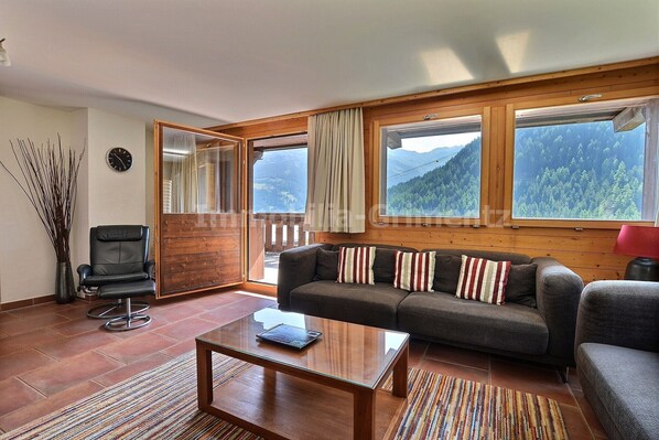 Ein Wohnzimmer mit einer Couch, einem Couchtisch, einem Sessel und großen Fenstern mit Blick auf die Berge.