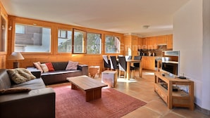 A cozy living room with a sofa, a coffee table, and a TV on a wooden stand. There is a dining area with a table and chairs. The room has wooden walls and large windows. - Very comfortable flat for 7 people, living room with wood-burner, kitchen area with dishwasher, 2 do (Grimentz)