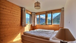 A bedroom with a wooden floor, a bed with white bedding, a lamp, and a window with curtains. - Very comfortable flat for 7 people, living room with wood-burner, kitchen area with dishwasher, 2 do (Grimentz)